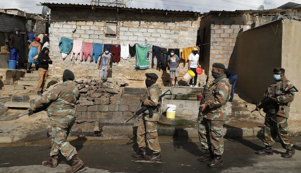 Orang-orang berdiri dekat rumah mereka dan menyaksikan anggota Pasukan Pertahanan Afrika Selatan (SANDF) berpatroli di daerah mereka di Alexandra, Johannesburg, Kamis (15/7/2021). Patroli tersebut menyusul kerusuhan massa yang disebabkan dipenjaranya mantan Presiden Jacob Zuma. (Phill Magakoe/AFP)