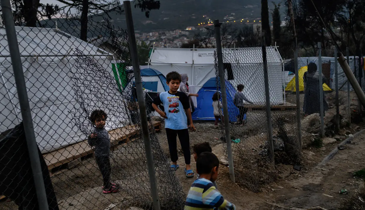Anak-anak imigran bermain di sebelah pagar di luar kamp pengungsi di pulau Samos, Yunani (13/11/2019). Kamp Samos, awalnya dibangun untuk menampung 650 orang, telah lama melampaui batas-batasnya. (AFP Photo/Angelos Tzortzinis)
