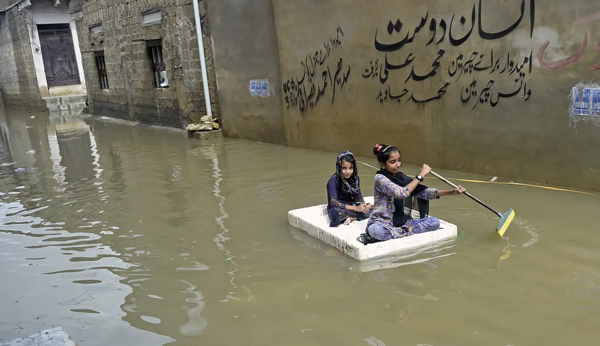 Gadis-gadis menggunakan rakit sementara melintasi jalan yang banjir di daerah perumahan setelah hujan muson lebat di Karachi (26/7/2022). Keadaan darurat cuaca diumumkan di Karachi karena hujan muson yang lebih deras dari biasanya terus melanda kota terbesar di Pakistan, membanjiri rumah-rumah dan membuat jalan-jalan tidak dapat dilalui. (AFP/Rizwan Tabassum)
