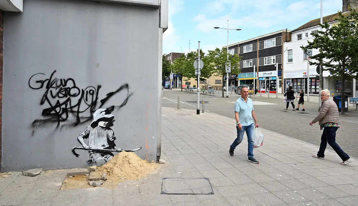 Pejalan kaki melewati stensil seorang anak menggali di pasir, yang menampilkan ciri-ciri seniman jalanan Banksy di dinding di Lowestoft di pantai timur Inggris pada 8 Agustus 2021. Karya tersebut belum diklaim oleh Banksy. (JUSTIN TALLIS / AFP)