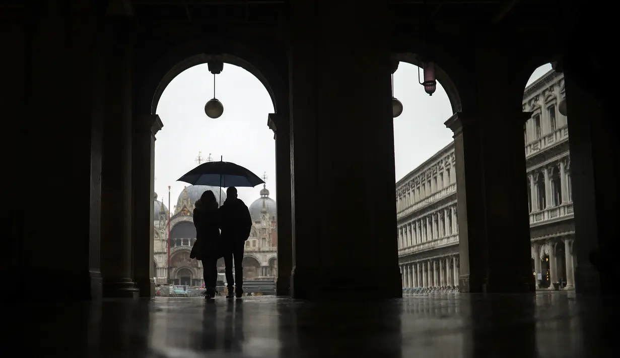 Suami istri mengenakan payung saat hujan di St. Mark's Square di Venesia, Minggu (1/3/2020). Pemerintah Italia akan melarang orang berciuman di tempat umum, berjabatan tangan dan menjaga jarak yang aman dari masing-masing lain untuk membatasi penyebaran virus corona. (AP Photo/Francisco Seco)