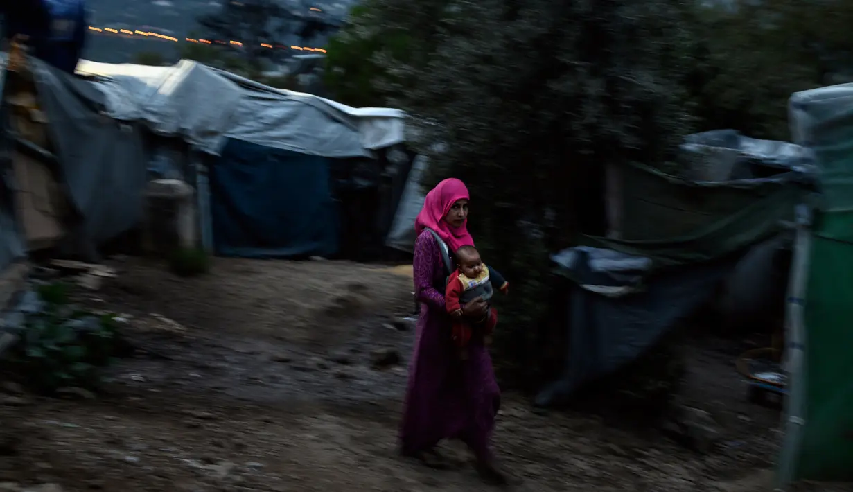Seorang wanita imigran menggendong bayinya berjalan di luar kamp pengungsi di pulau Samsos, Yunani (13/11/2019). Ribuan imigran berjuang untuk menemukan ruang di perbukitan di atas kota, membuat rumah sementara dari bahan bangunan apa pun yang dapat mereka temukan. (AFP Photo/Angelos Tzortzinis)