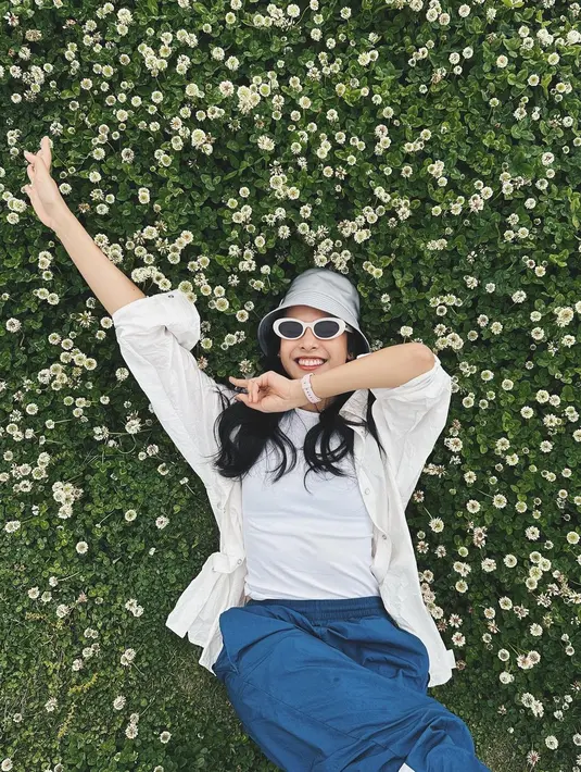 Maudy Ayunda terlihat cantik mengenakan busana kasual. Dalam foto ini, Maudy Ayunda berbaring di rumput dengan bunga-bunga putih mengenakan kaus putih, dilapisi dengan kemeja lengan panjang yang juga berwarna putih, dipadu dengan celana biru, bucket hat, dan sunglasses. Foto: Instagram.