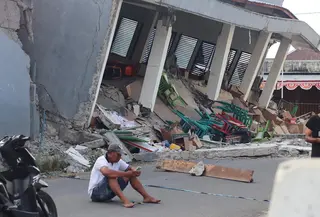Seorang pria memeriksa ponselnya di dekat bangunan yang runtuh akibat gempa bumi di Reok, Provinsi Nusa Tenggara Timur, Indonesia, Minggu, 16 Agustus 2026. (Foto AP/Aquila Handayani Abbas)