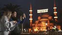 Dua wanita berdiri di dekat Masjid Sultan Ahmed yang ikonik, lebih dikenal sebagai Masjid Biru, dihiasi dengan lampu dan slogan bertuliskan "Ramadhan adalah cinta," menandai bulan Ramadhan, di distrik bersejarah Sultan Ahmed di Istanbul (13/4/2021). (AP Photo/Emrah Gurel)