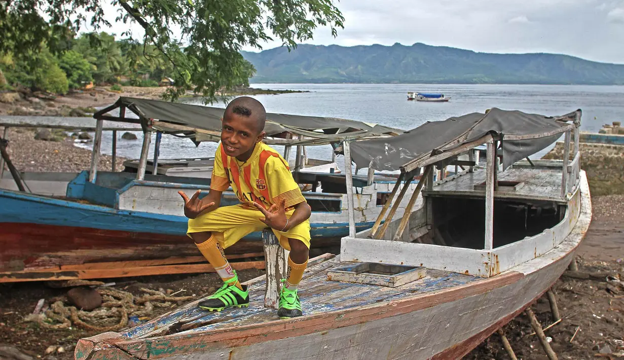 Seorang Anak duduk diatas perahu dengan jersey Barcelona siap untuk bermain sepak bola di Alor, Nusa Tenggara Timur. (Bola.com/Nicklas Hanoatubun)