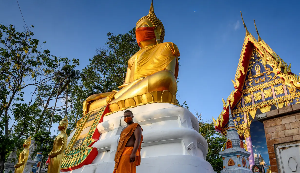 Biksu berjalan di depan patung Buddha raksasa yang mengenakan masker di kuil Wat Nithet Rat Pradit di Pathum Thani di luar Bangkok, Thailand, 12 Mei 2020. Pemasangan masker tersebut sebagai tanggapan terhadap penyebaran pandemi Covid-19. (Photo by Mladen ANTONOV / AFP)