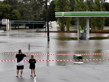 Sebuah keluarga berjalan mengunjungi pompa bensin yang terendam banjir di pinggiran barat daya Camden, Sydney, Australia, Selasa (8/3/2022). (Muhammad FAROOQ/AFP)