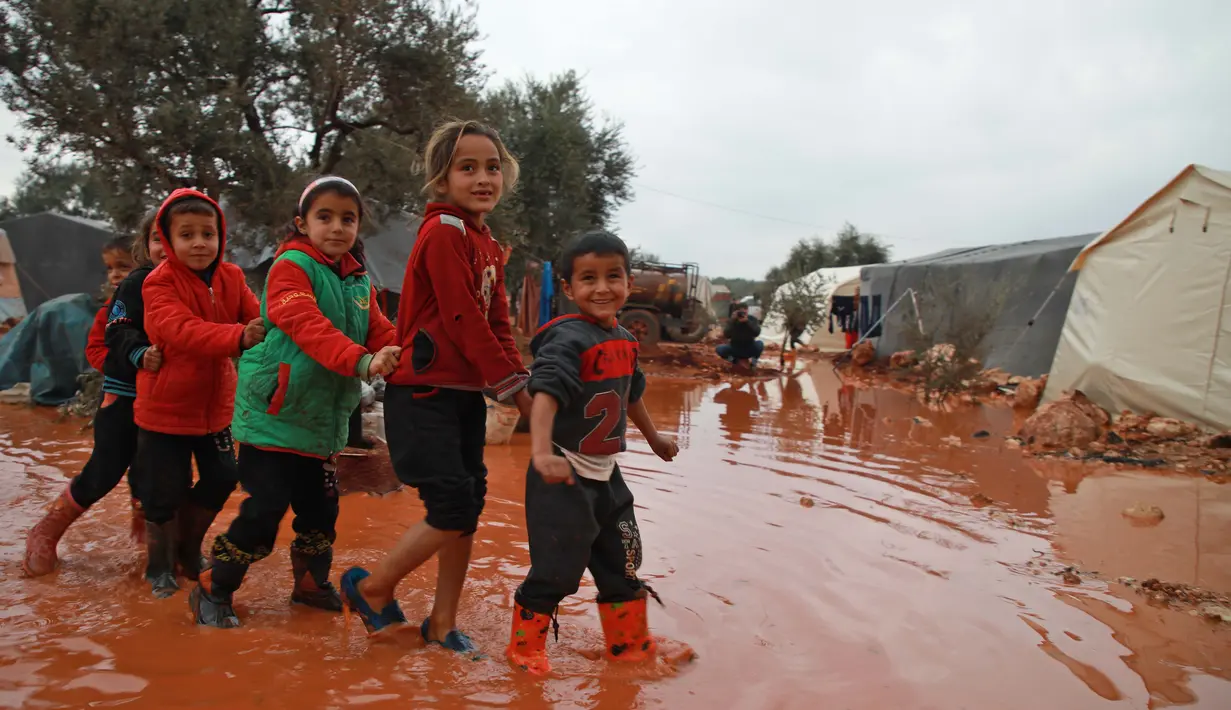 Anak-anak berjalan melewati genangan air hujan di depan tenda di sebuah kamp untuk warga Suriah yang kebanjiran di dekat desa Killi di utara provinsi Idlib barat laut (5/12/2019). (AFP Photo/Aaref Watad)