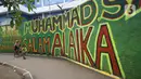 Warga berjalan di depan mural bernuansa Islami di Gang Pelangi, kawasan Kalibata, Jakarta, Jumat (23/4/2021). Mural bernuansa Islami tersebut dibuat untuk menghiasi dan meramaikan pintu masuk gang sekaligus mengingatkan masyarakat untuk selalu beribadah dan bershalawat (Liputan6.com/Herman Zakharia)