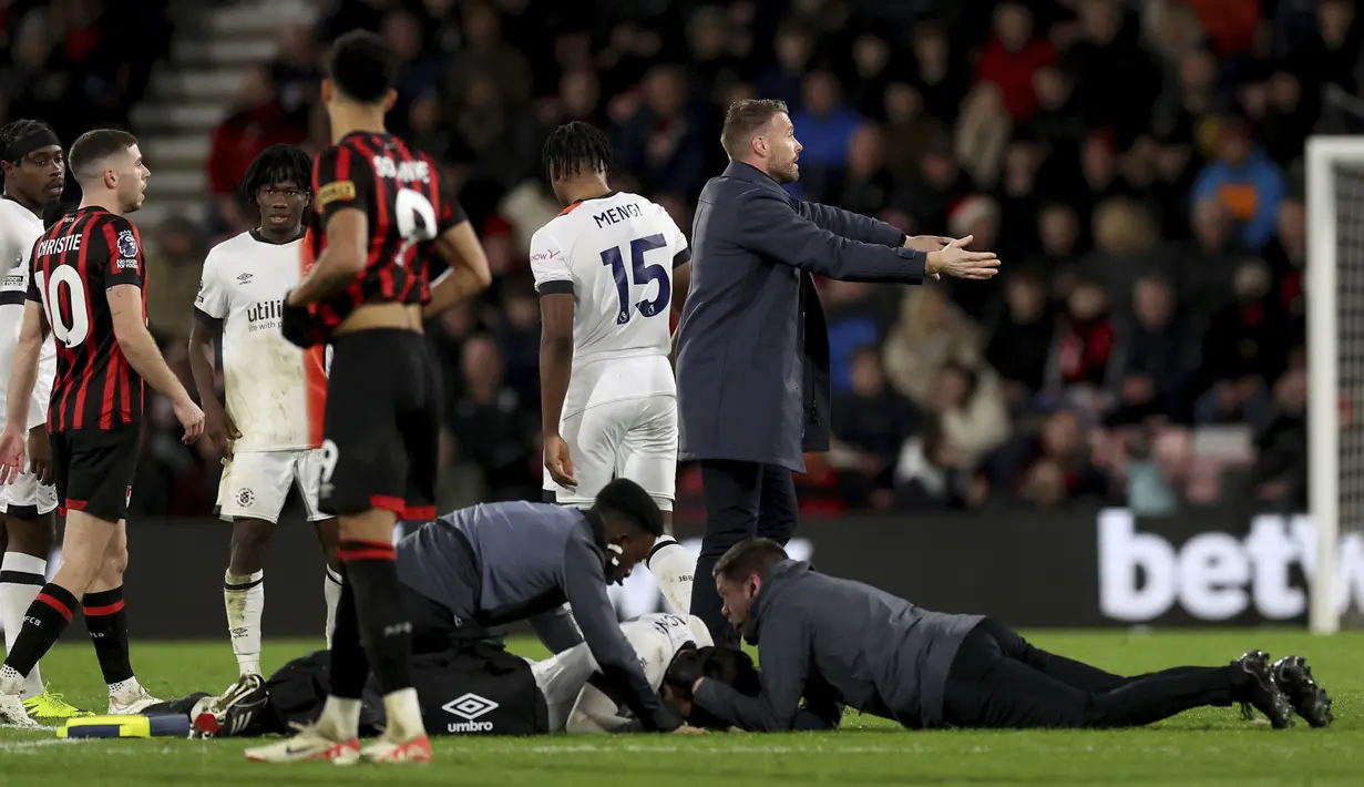 <p>Pelatih Luton Town, Rob Edwards (kanan) menenangkan para pemain saat petugas medis menolong Tom Lockyear yang mengalami pingsan secara tiba-tiba di tengah lapangan dalam pertandingan Liga Inggris 2023/2024 antara Bournemouth melawan Luton Town yang berlangsung di Vitality Stadium, Sabtu (16/12/2023). (AP/PA/Steven Paston)</p>