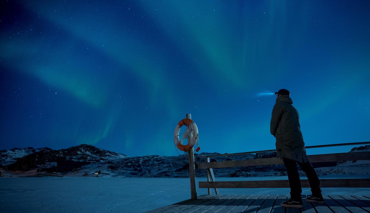 Seorang pria berdiri di dermaga untuk menonton Aurora Borealis atau Northern Lights di Kepulauan Lofoten, Bostad, Arktik, Norwegia, Sabtu (3/3). Aurora menampilkan banyak warna, tapi yang sering muncul adalah hijau muda dan pink. (Olivier MORIN/AFP)