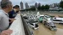 Orang-orang melihat mobil-mobil yang terendam banjir setelah hujan lebat melanda kota Zhengzhou di provinsi Henan, China tengah (21/7/2021). Luapan sungai menggenangi jalan-jalan dan membuat kendaraan terbawa arus setelah curah hujan 200 mm turun dalam satu jam. (AFP/STR)