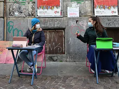 Anita Iacovelli (kiri) dan temannya Lisa Rogliatti, berusia 12 tahun, duduk di depan sekolah Italo Calvino di Turin, Italia, 17 November 2020. Anita memprotes penutupan sekolahnya karena pembatasan COVID-19 dengan duduk di luar gedung sambil mengikuti pelajaran jarak jauh. (Miguel MEDINA/AFP)