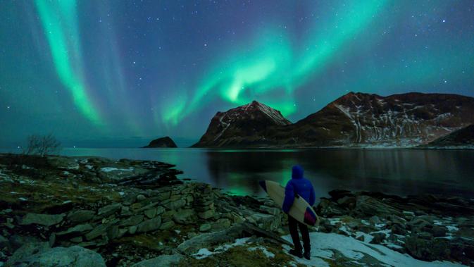 Seorang surfer melihat Cahaya Utara atau aurora borealis di Utakleiv, Norwegia utara (9/3). Cahaya kutub terbentuk dari interaksi medan magnet Bumi dengan lapisan terluar Matahari atau Korona. (AFP/Olivier Morin)