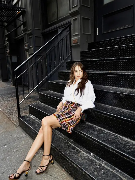 Gaya Enzy Storia yang memukau seperti biasanya duduk berpose di tangga. Ia mengenakan kemeja putih lengan panjang, dipadu rok kotak-kotak beraneka warna, dipadu sling mini bag hitam, dan open toe heels yang juga berwarna hitam. [Foto: Instagram/enzystoria]