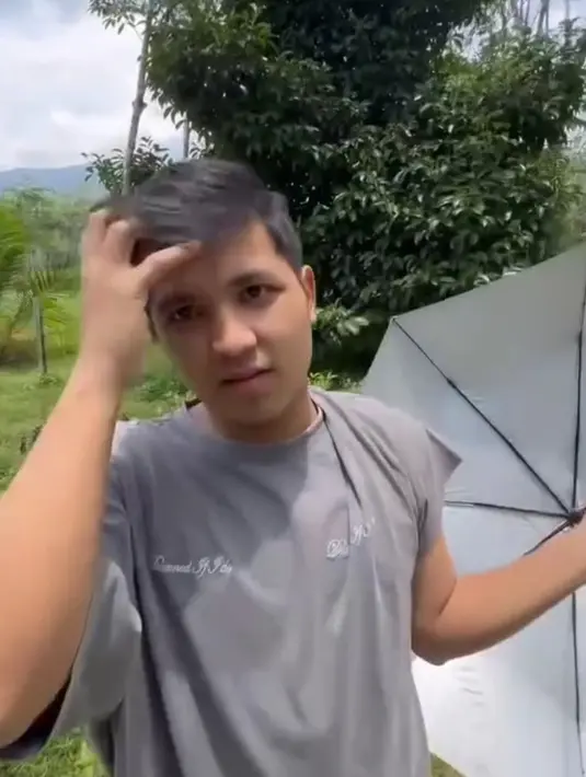 Penampilannya begitu sederhana dengan kaos abu-abu longgar dan celana hitam santai. Sambil berjalan di antara pepohonan karet, Jirayut menunjukkan bagaimana proses penyadapan getah dilakukan. Sesekali, ia bercanda dengan menunjukkan wadah kecil berisi getah karet, membuat suasana terasa akrab dan ceria. (Liputan6.com/IG/@jirayutdaa4official)