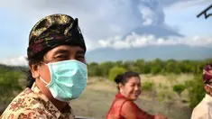 Seorang pria mengenakan masker saat menaiki mobil bak terbuka melewati Gunung Agung di Kabupaten Karangasem, Bali (27/11). Sebelumnya, Gunung Agung telah mengalami erupsi sejak Sabtu (25/11) sore.  (AFP/Sonny Tumbelaka)
