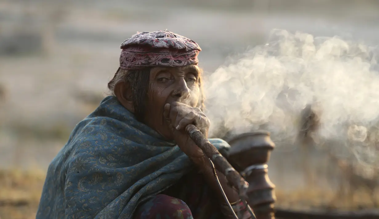 Seorang wanita nomaden merokok hukka, atau gelembung hubble, saat kelompok mereka berhenti sementara di jalan raya Jammu-Srinagar di Nagrota, di pinggiran Jammu, India, Selasa (9/11/2021). Anggota suku tersebut pindah dari perbukitan untuk menghindari musim dingin. (AP Photo/Channi Anand)