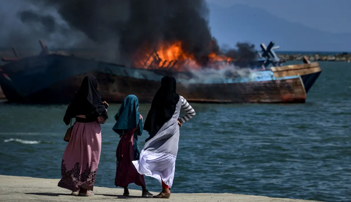 Para warga menyaksikan asap hitam mengepul dari kapal asing ilegal yang dibakar dan ditenggelamkan Kejaksaan Negeri Banda Aceh di perairan Pelabuhan Perikanan Samudera Kutaraja, Banda Aceh, Aceh, Kamis (18/3/2021). (AFP/Chaideer Mahyuddin)