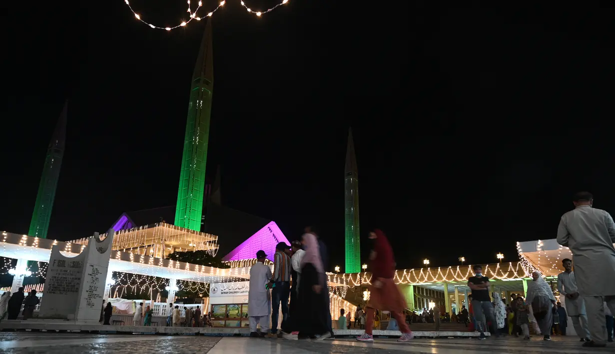 Orang-orang melewati bangunan masjid Faisal yang diterangi selama perayaan hari kelahiran Nabi Muhammad SAW atau Maulid Nabi di Islamabad, Pakistan, Senin (18/10/2021). Peringatan Maulid Muhammad tak hanya diperingati umat Islam di Indonesia, tapi juga di berbagai negara. (Farooq NAEEM/AFP)