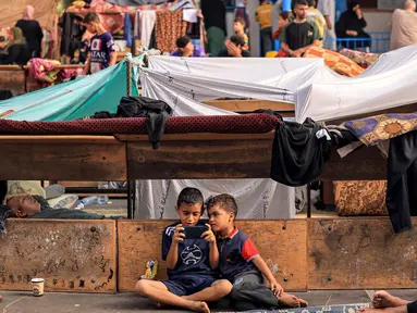 Seorang anak duduk di samping anak lainnya yang sedang bermain ponsel di taman bermain sekolah yang dikelola oleh Badan Bantuan dan Pekerjaan PBB untuk Pengungsi Palestina (UNRWA), 25 Oktober 2023. (MAHMUD HAMS/AFP)