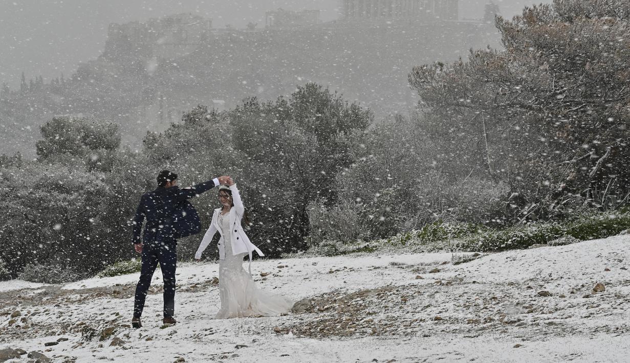Pasangan pengantin baru dari AS menikmati hujan salju selama pemotretan di dekat Acropolis di Athena, Senin (24/1/2022). Sekolah-sekolah ditutup di daerah Athena terbesar, lantaran Yunani mengalami badai musim dingin dengan hujan salju lebat dan suhu yang sangat rendah. (Louisa GOULIAMAKI/AFP)
