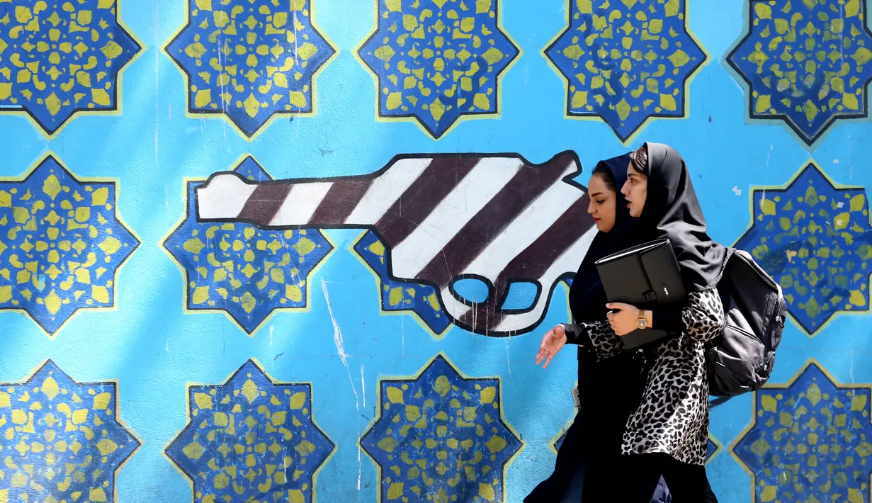 Dua orang wanita berjalan melewati lukisan mural yang menggambarkan pistol di sepanjang dinding bekas Kedutaan Amerika Serikat (AS) di Ibu Kota Teheran, Iran, Sabtu (22/6/2019). (ATTA KENARE/AFP)