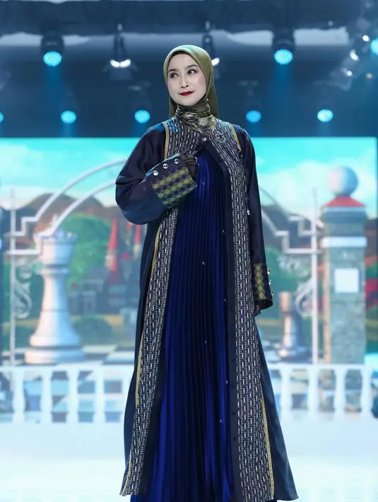 Desy Ratnasari tampil memancarkan aura elegan yang tenang dalam balutan busana bernuansa gelap, perpaduan hitam dan biru navy yang terasa sophisticated. Siluetnya dibuat lurus dan memanjang, dengan inner dress bertekstur pleats halus berwarna biru tua yang memberi efek jatuh ringan setiap kali ia melangkah. Di bagian luar, ia mengenakan outer panjang berpotongan clean dengan panel bordir vertikal bernuansa emas-keperakan. [Dok/Shi by Shireen & Zaskia Sungkar].