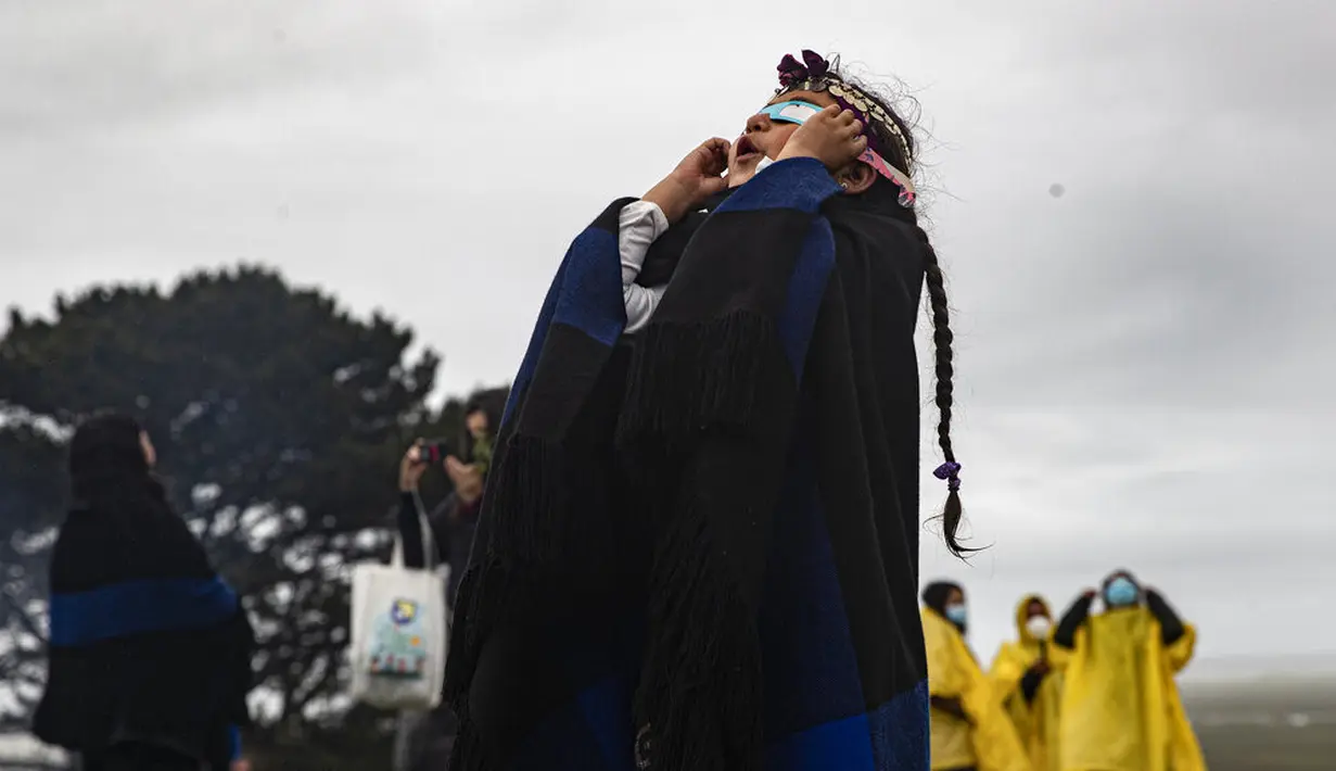 Magdalena Nahuelpan, gadis Adat Mapuche, melihat gerhana matahari total melalui langit mendung menggunakan kertas khusus dan gelas plastik di Carahue, La Araucania, Chile, Senin (14/12/2020). Gerhana matahari total terlihat dari Chile dan wilayah Patagonia utara Argentina. (AP Photo/Esteban Felix)