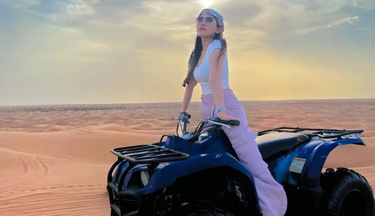 Menaiki ATV sambil berpose berdiri. Terlihat Via mengenakan celana panjang warna ungu, kaos ketat warna putih, sorban, dan kacamata hitam. (Instagram/viavallen)
