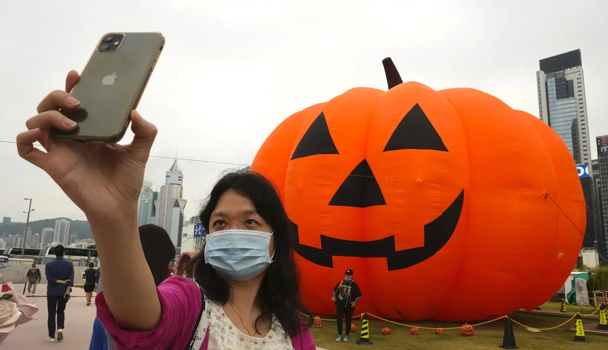 Seorang wanita mengenakan masker untuk mencegah penyebaran virus corona, berpose untuk selfie di depan dekorasi buah labu besar untuk Halloween di sebuah taman di Hong Kong, Rabu (27/10/2021). Tahun ini, perayaan tersebut jatuh pada hari Minggu, 31 Oktober 2021. (AP Photo/Vincent Yu)