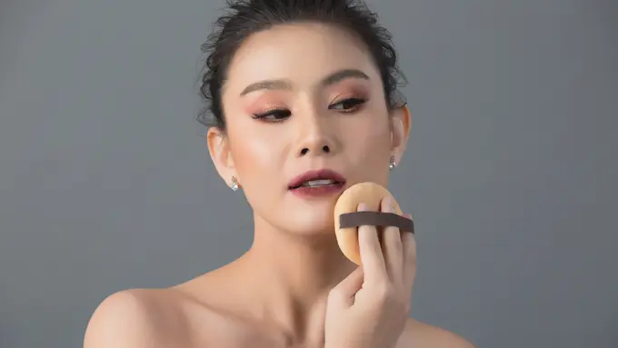 Seorang perempuan sedang mengaplikasikan bedak menggunakan puff ke wajahnya (Foto Dok: Freepik/jcomp)
