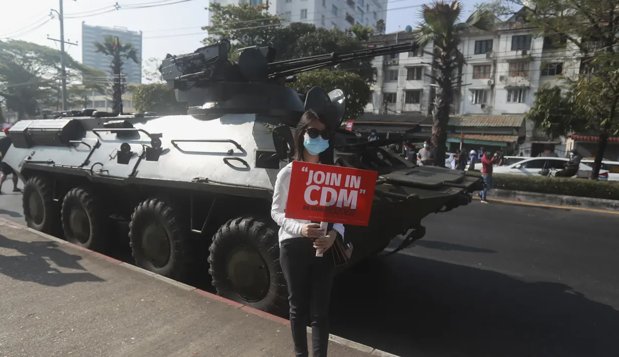 Seorang pengunjuk rasa anti-kudeta memegang poster saat berpose di depan kendaraan lapis baja yang dikerahkan di luar gedung Bank Sentral Myanmar di Yangon, Senin (15/2/2021). Pasukan militer Myanmar mulai menerjunkan kendaraan lapis baja di kota-kota besar dan memutus akses internet. (AP Photo)