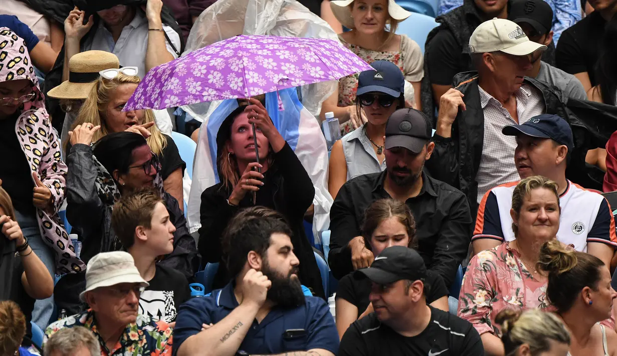 Seorang wanita berlindung di bawah payung saat hujan selama pertandingan tunggal putaran pertama antara Roger Federer dari Swiss dan Steve Johnson dari Amerika Serikat di kejuaraan tenis Australia Terbuka di Melbourne, Australia, Senin (20/1/2020). (AFP Photo/William West)