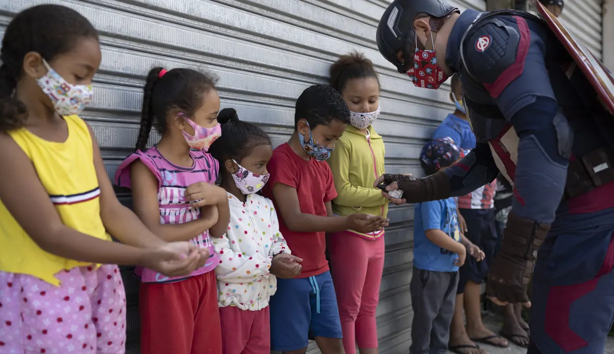 Polisi militer Everaldo Pinto, berkostum Captain America, menyemprotkan cairan pembersih ke tangan anak-anak di Petropolis, Brasil, Kamis (15/4/2021). Dalam aksinya, Pinto membagikan kotak berisi produk pembersih dan masker untuk mencegah paparan corona pada anak-anak. (AP Photo/Silvia Izquierdo)