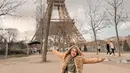 Berpose di depan menara Eiffel, Ria Ricis tampil kece memadukan gamis dan hijab nuansa hijau sage-nya dengan long coat warna coklat muda. [@riaricis1795]