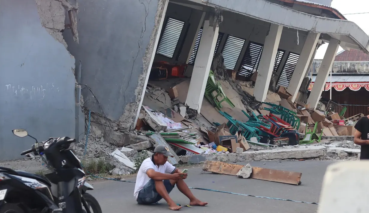 Seorang pria memeriksa ponselnya di dekat bangunan yang runtuh akibat gempa bumi di Reok, Provinsi Nusa Tenggara Timur, Indonesia, Minggu, 16 Agustus 2026. (Foto AP/Aquila Handayani Abbas)