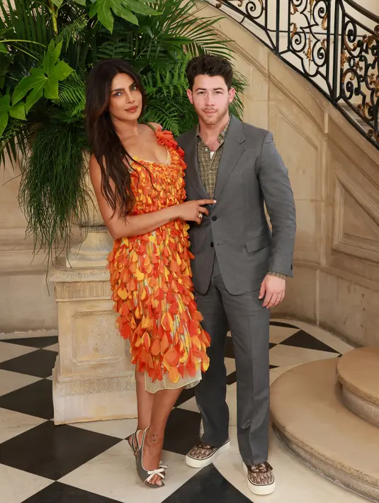 Priyanka Chopra dan Nick Jonas hadiri Dior Haute Couture dengan busana yang kontras. Priyanka kenakan v-neck dress full petal bernuansa oranye dan Nick Jonas kenakan setelan abu-abu dan kemeja kotak-kota [Dior]