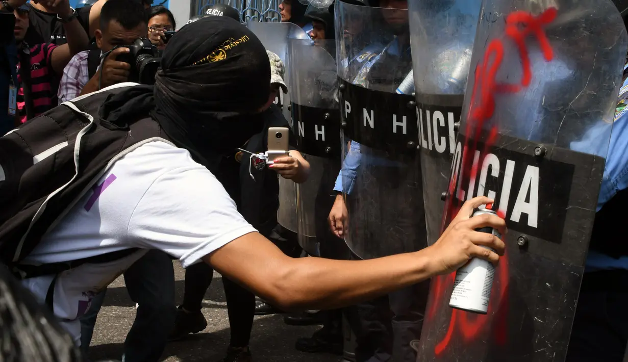 Seorang demonstran menyemprotkan cat di tameng polisi anti huru hara saat melakukan aksi protes pada peringatan Hari Buruh di Tegucigalpa, Honduras (1/5). (AFP/Orlando Sierra)