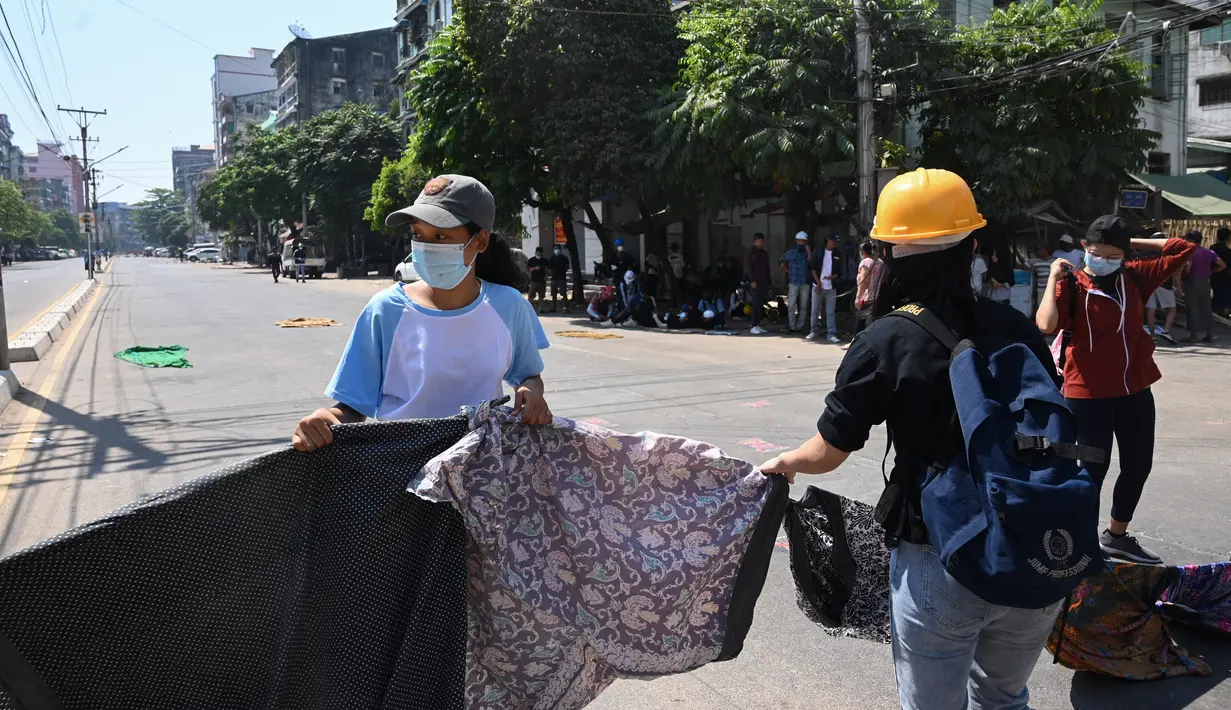 Perempuan menggantung pakaian tradisional Myanmar, longyi, di seberang jalan selama demonstrasi menentang kudeta militer di Yangon, Senin (8/3/2021). Jemuran kain itu untuk membatasi gerak polisi dan tentara karena berjalan di bawah jemuran pakaian ini dianggap akan membawa sial bagi pria. (STR/AFP)