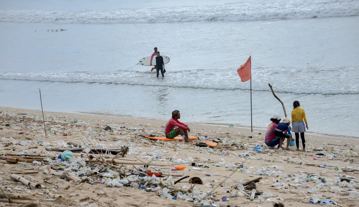Orang-orang duduk di antara sampah kiriman yang terdampar memenuhi pesisir pantai Kuta, Bali, Kamis (31/12/2020). Menjelang pergantian tahun baru, Pantai Kuta hanya terlihat beberapa wisatawan, namun tumpukan sampah kiriman tersebar di sepanjang bibir pantai. (SONNY TUMBELAKA / AFP)