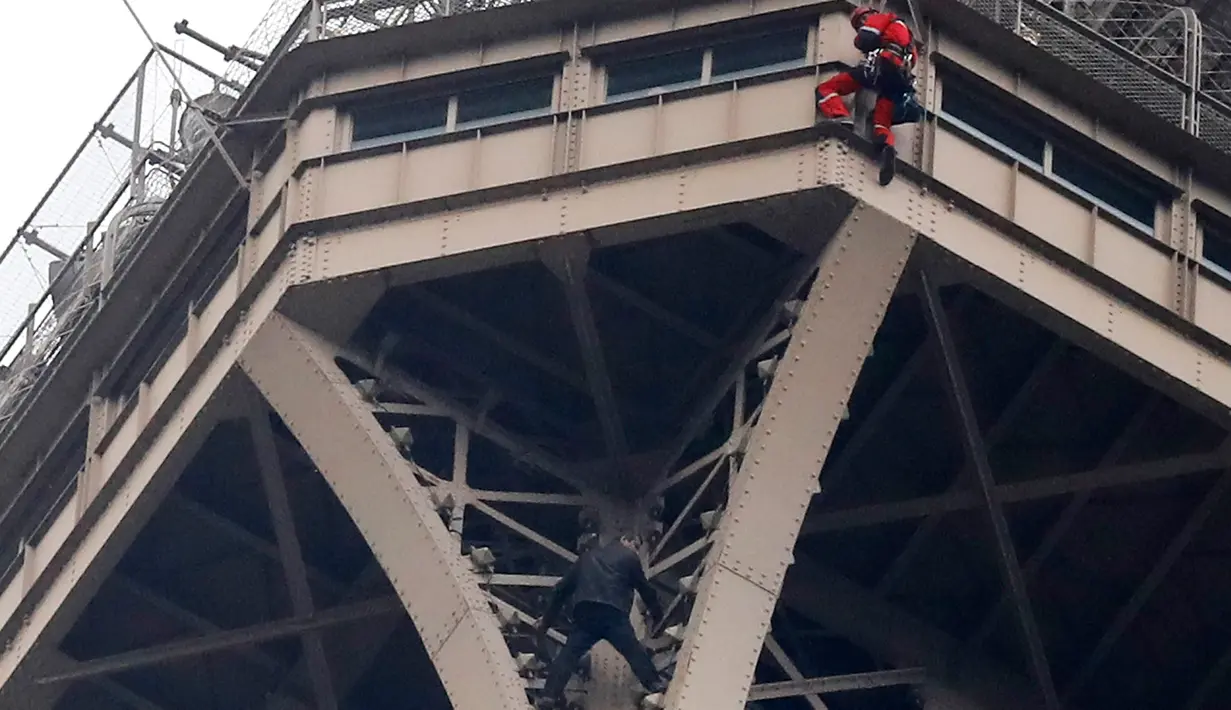 Petugas pemadam kebakaran berusaha mendekati seorang pria (baju hitam di bawah) yang memanjat Menara Eiffel tanpa peralatan keselamatan di Paris, Senin (20/5/2019). Akibat ulahnya, Menara Eiffel ditutup dan pihak pengelola melakukan evakuasi dan pengamanan. (AP/Michel Euler)
