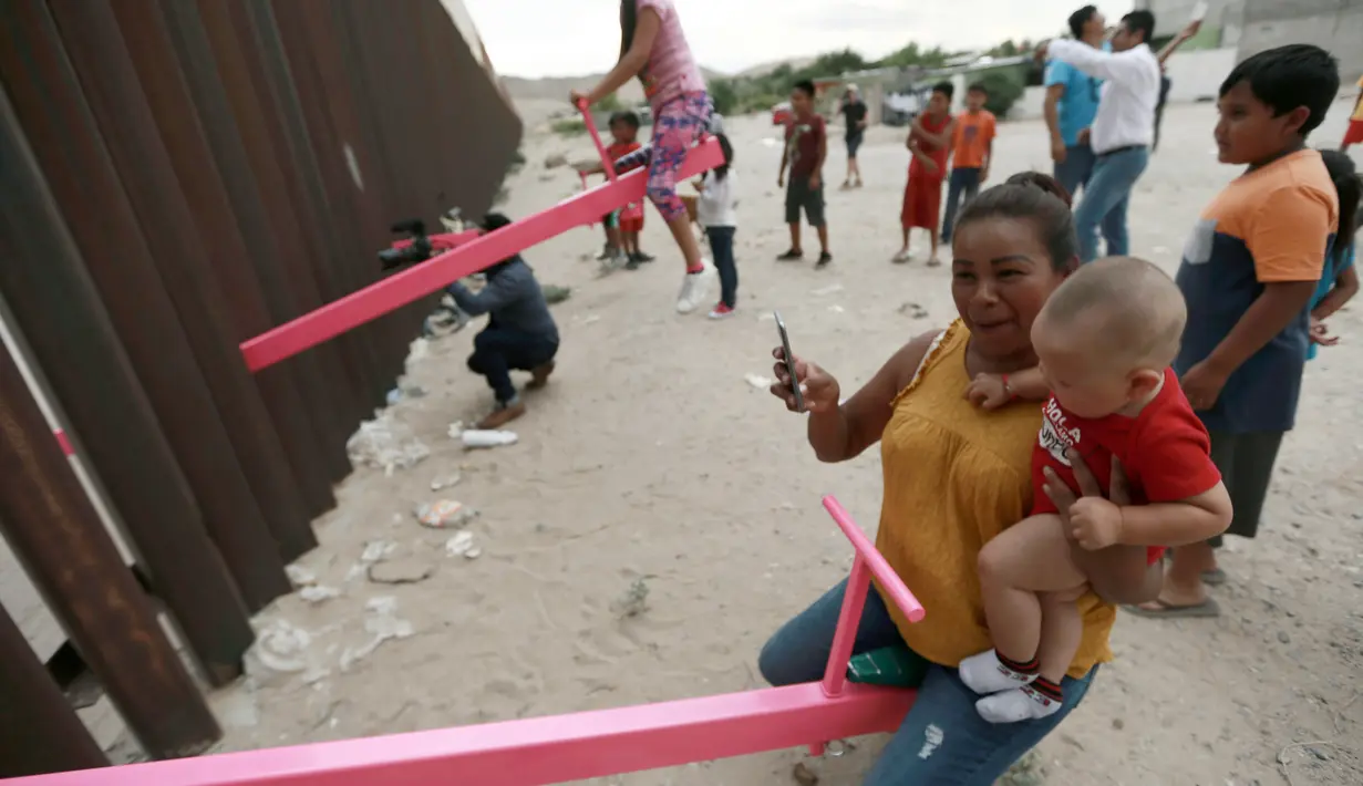 Seorang ibu dan bayinya bermain jungkat-jungkit yang dipasang di antara pagar pemisah Meksiko dengan Amerika Serikat, Ciudad de Juarez, Meksiko, Minggu (28/7/2019). Jungkat-jungkit tersebut dirancang seorang profesor arsitektur California, Ronald Rael. (AP Photo/Christian Chavez)