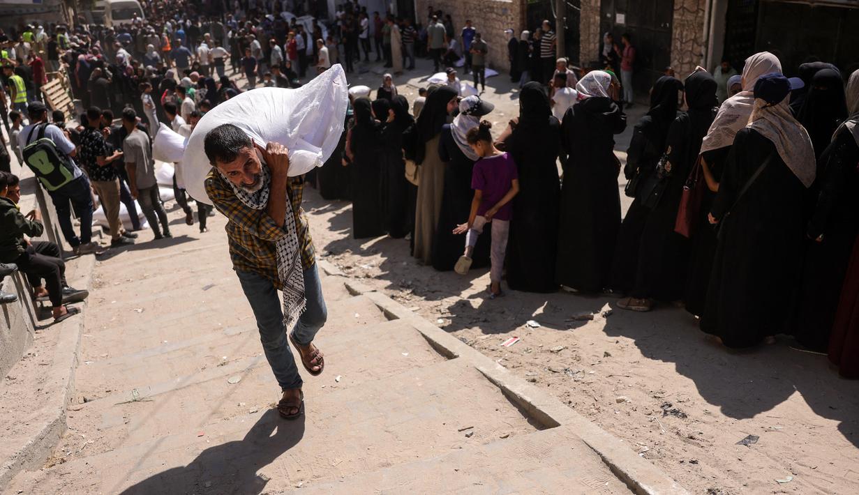 Seorang pria Palestina membawa sekantong tepung, sementara yang lain menunggu di depan titik distribusi yang didirikan oleh Save Youth Future Society (SYFS), yang bekerja sama dengan Program Pangan Dunia PBB (WFP), di Kota Gaza pada 26 Juni 2025. (BASHAR TALEB/AFP)