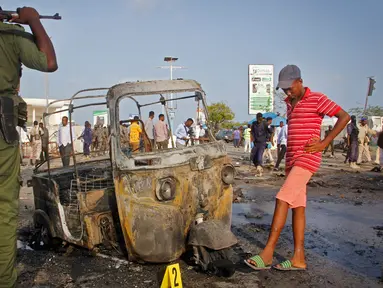 Tentara dan seorang anak berdiri dekat puing-puing kendaraan di lokasi ledakan bom mobil dekat pos pemeriksaan parlemen dan Kementerian Dalam Negeri di Ibu Kota Somalia, Mogadishu, Minggu (25/3). Serangan menewaskan empat orang. (AP/Farah Abdi Warsameh)