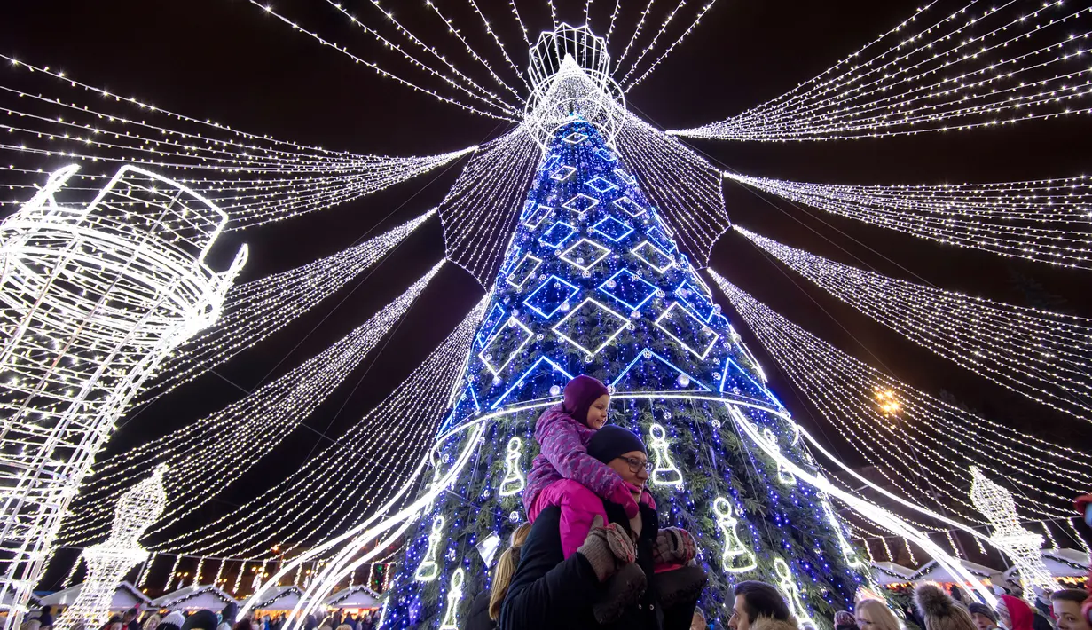Orang-orang berjalan di dekat pohon Natal raksasa yang menerangi Alun-Alun Katedral di Vilnius, Lithuania, Minggu (1/12/2019). Secara tradisi, pohon Natal Vilnius biasanya berupa rangka logam yang ditutupi dengan dekorasi dan cabang-cabang pohon dari hutan. (AP/Mindaugas Kulbis)