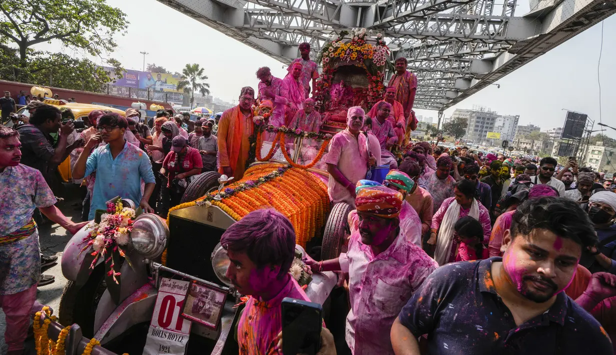 Umat Hindu berkerumun di sekitar mobil Rolls Royce 1921 yang dimodifikasi membawa patung dewa Krishna melalui bangunan bersejarah Howrah Bridge selama festival Holi di Kolkata, India, Minggu, 5 Maret 2023. (AP Photo/Bikas Das)