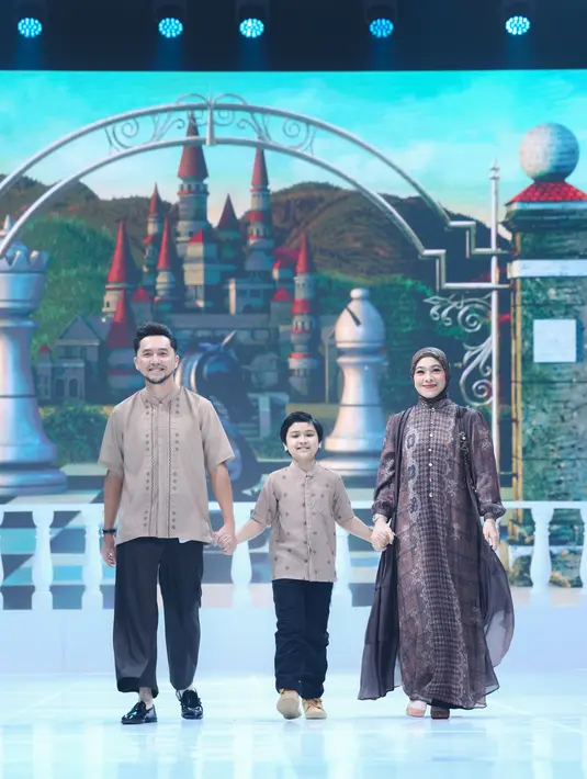 Diana Ayu Lestari tampil cerah dan penuh kehangatan bersama sang suami, Ananda Omesh, serta putra mereka dalam momen runway yang terasa sangat personal. Diana memilih long dress bermotif geometris bernuansa cokelat tua, marun, dan sentuhan ungu yang terlihat kaya tekstur. Siluetnya lurus dengan potongan ringan, diperkaya layering sheer di bagian bawah rok yang memberi efek flowy saat ia bergerak. Omesh tampil santai namun tetap polished dalam kemeja lengan pendek berwarna beige dengan detail bordir minimal di bagian depan. Sang putra tampil kompak di tengah, mengenakan atasan bernuansa cokelat muda dengan motif kecil yang selaras dengan palet busana orang tuanya.  [Dok/Shi by Shireen & Zaskia Sungkar].
