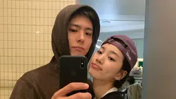 Suzy dan Park Bo Gum melakukan mirror selfie. Bo Gum mengenakan hoodie sementara Suzy berpenampilan boyish dengan hoodie dan topi.